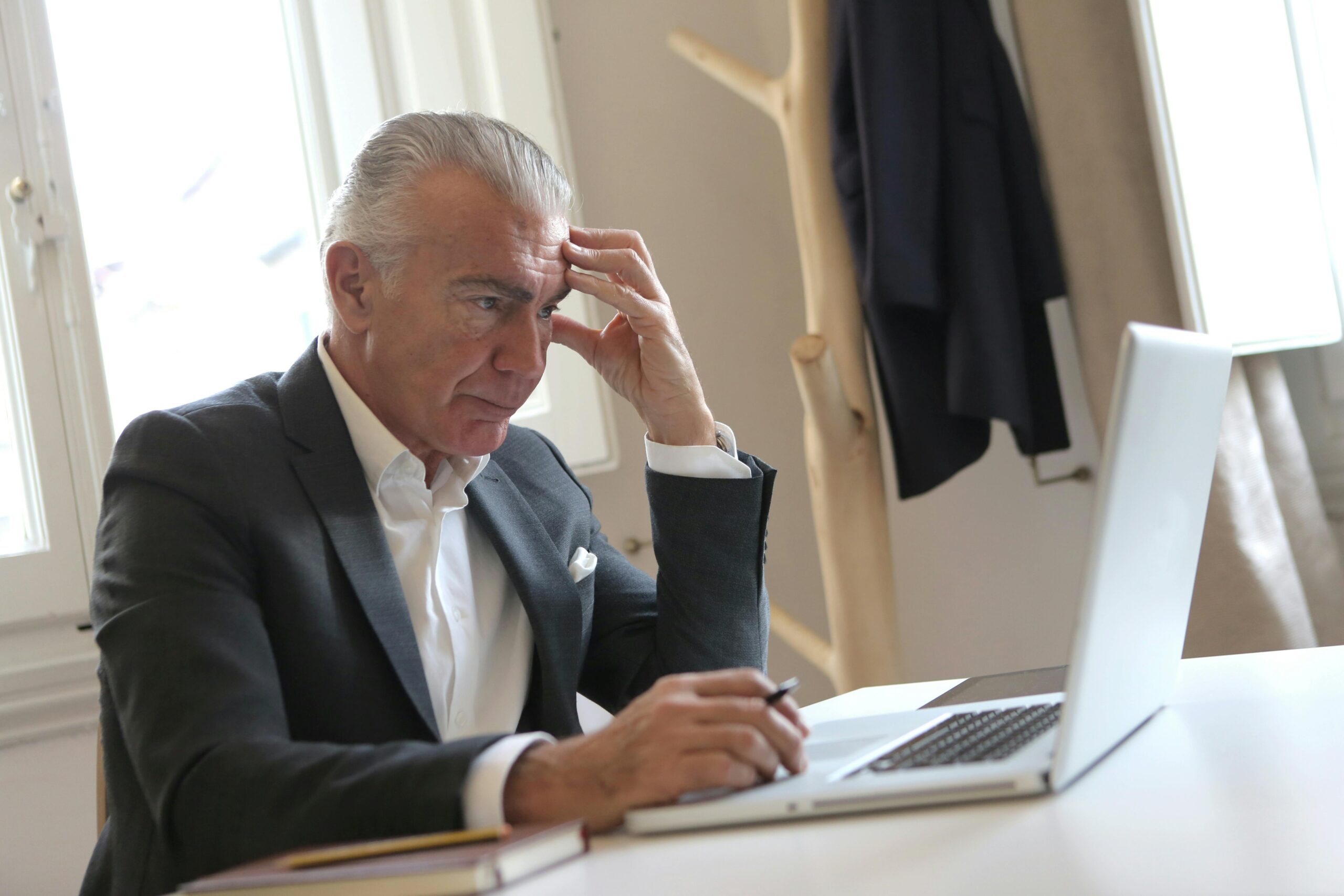 A stressed older man sitting with his hand on his temple while holding a pen and looking at his laptop | Source: Pexels