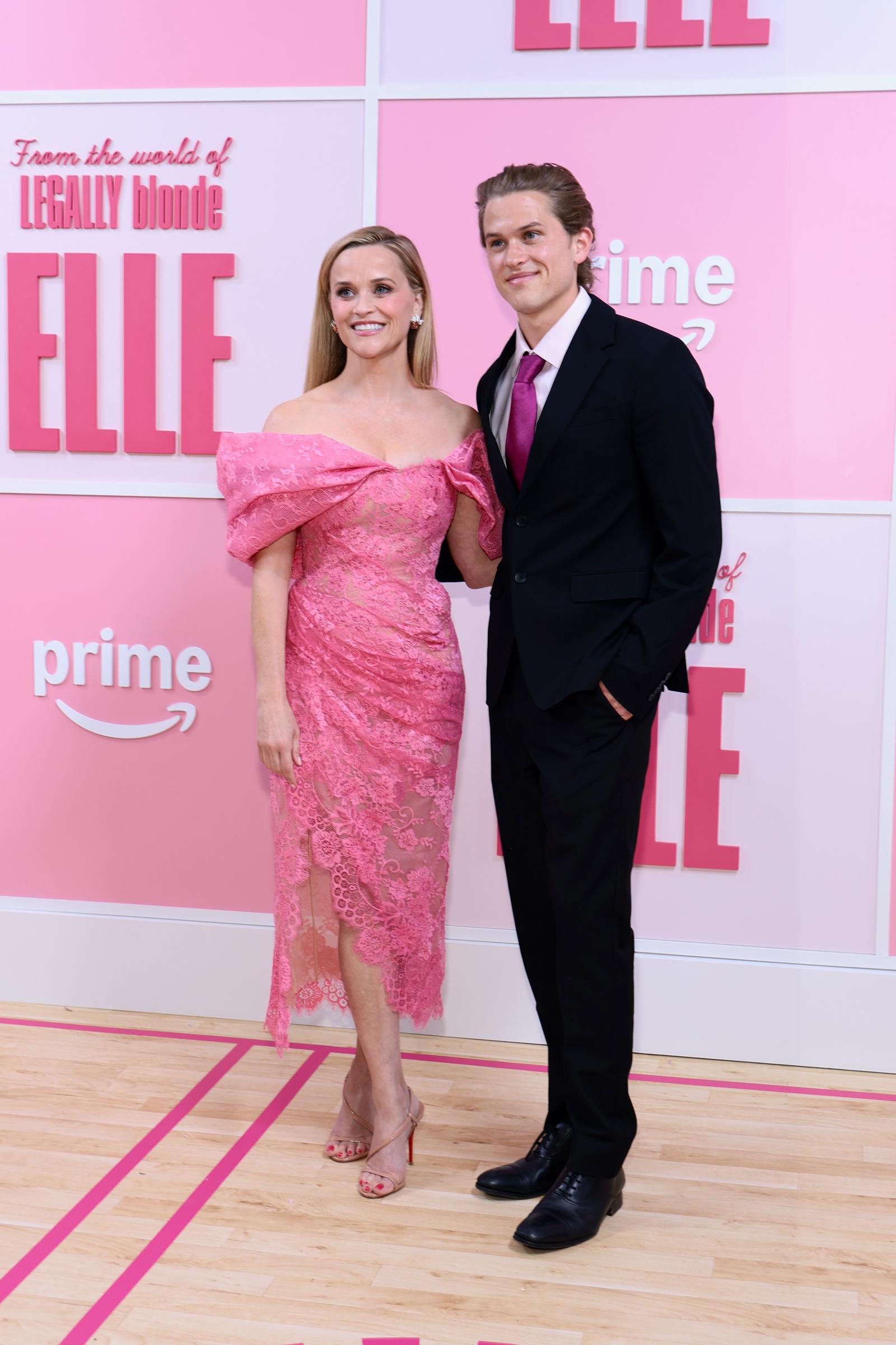 Reese Witherspoon and Deacon Phillippe pose at the 2026 "Elle" Season 1 world premiere in New York City, with her in a pink lace dress and him in a black suit with a magenta tie.