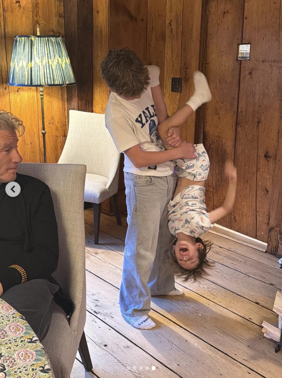 Richard Gere looks on as children play indoors in a cozy room featuring wood-paneled walls, rustic flooring, and soft lighting.