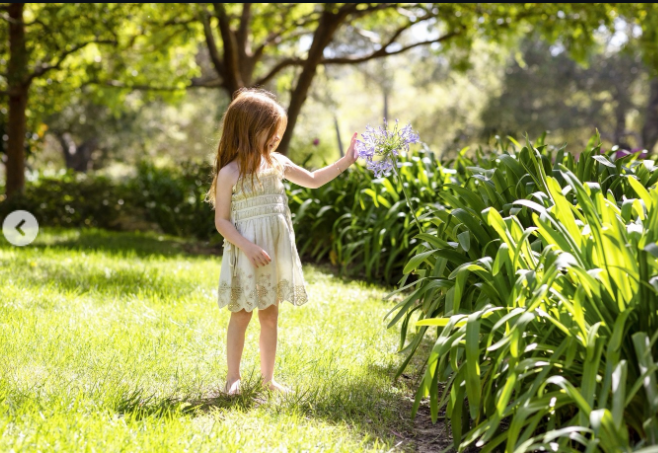 Princess Lilibet explores a sunlit garden while reaching toward a cluster of purple flowers, her cream-colored dress and long red hair standing out against the lush greenery.