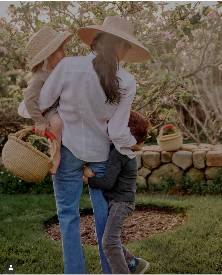 Meghan Markle walks through a blooming orchard carrying one child while another clings to her side, capturing a candid moment of family life outdoors.