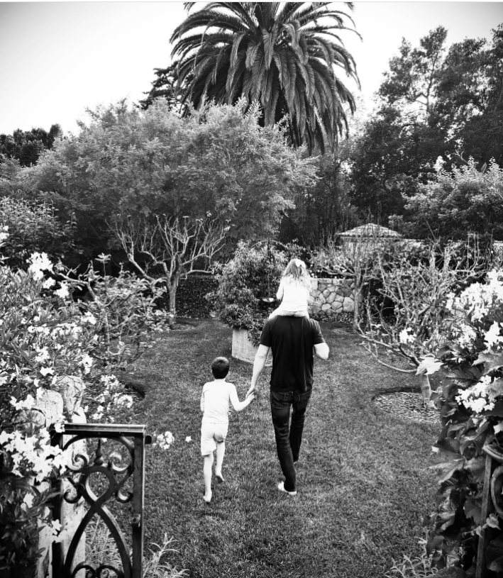 Prince Harry strolls through a manicured garden with his children, carrying Princess Lilibet on his shoulders while holding Prince Archie's hand.