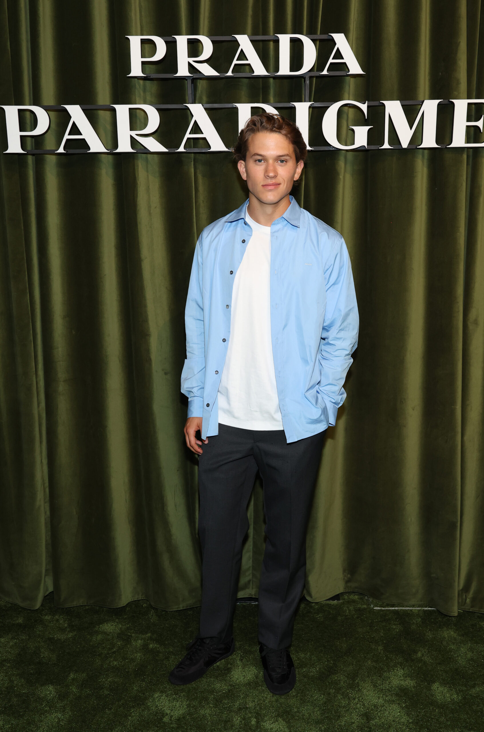 Deacon Reese Phillippe attends Prada's Paradigme Global Launch Party in New York City, posing solo in a pale blue shirt, white tee, dark pants, and black sneakers.