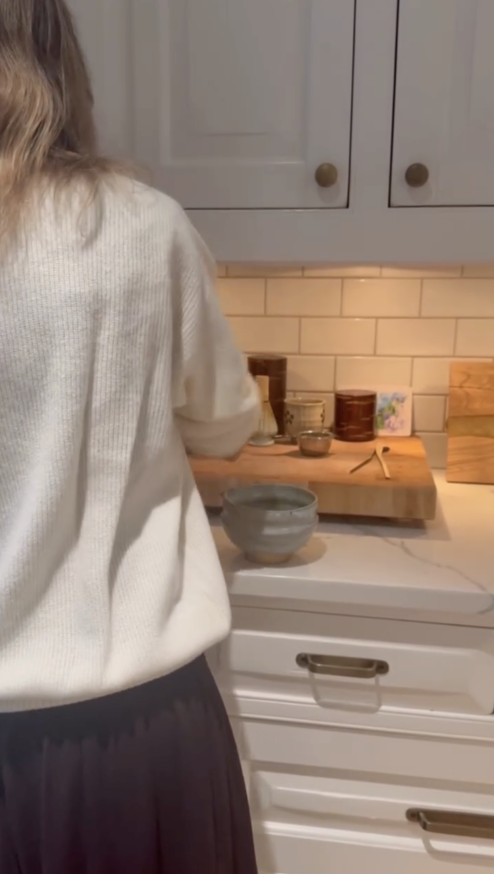 Alejandra Silva stands at a kitchen counter inside the family's Spain home, offering a glimpse of the bright space with white cabinetry, subway-tile backsplashes, and warm under-cabinet lighting.