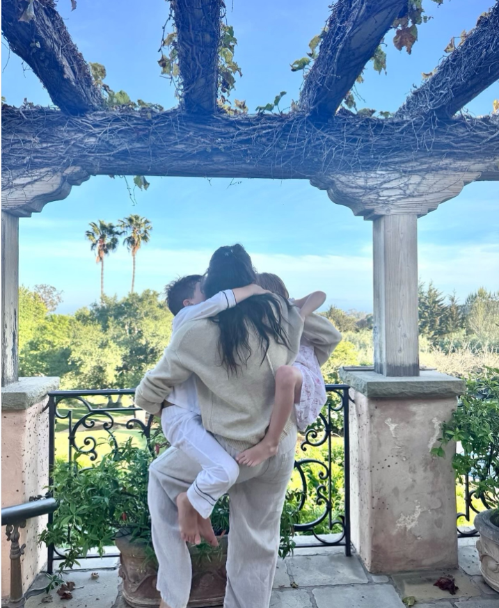 Meghan Markle stands beneath a vine-covered pergola while embracing Prince Archie and Princess Lilibet in a joyful family hug.
