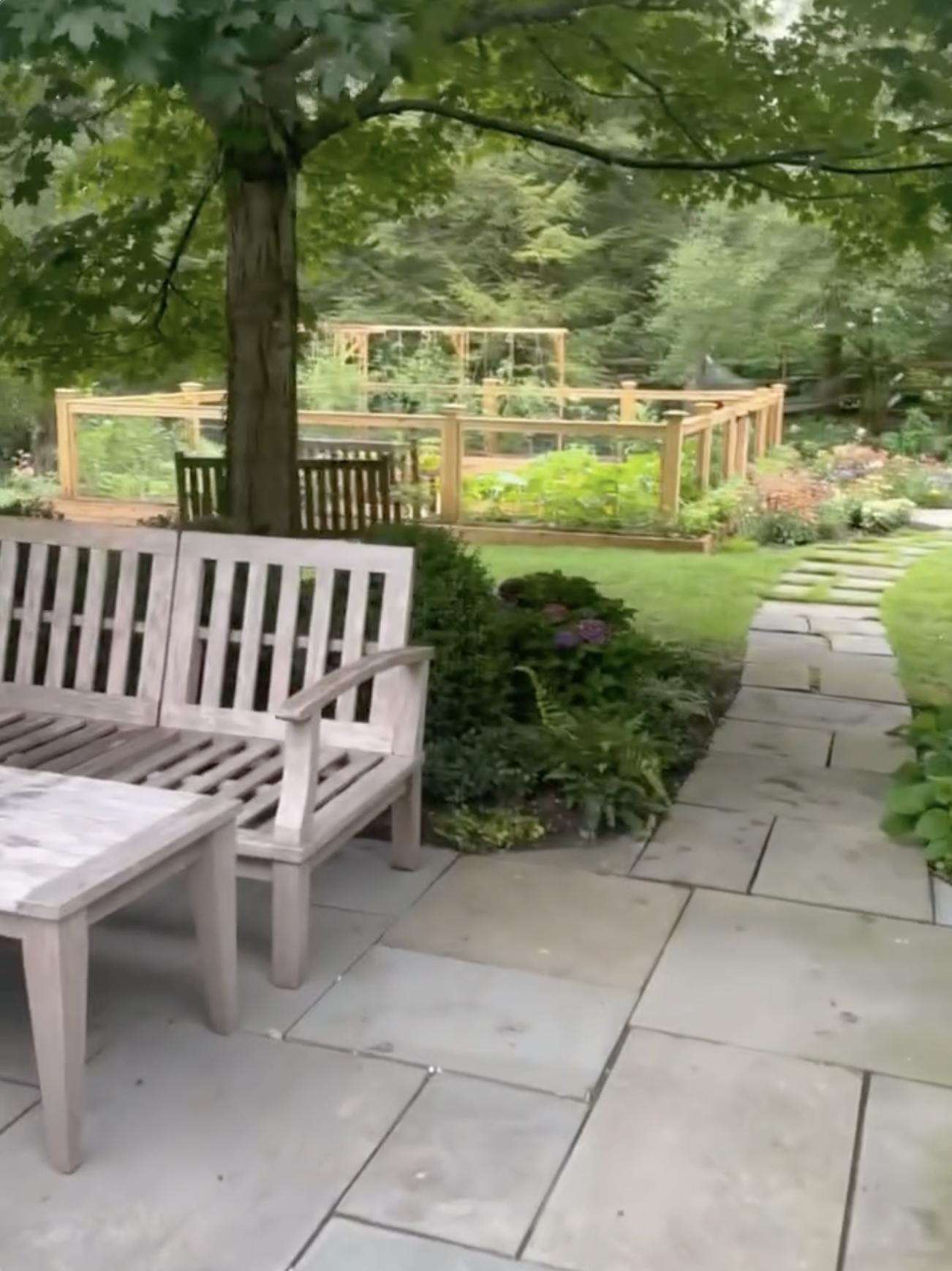 A winding pathway leads past outdoor seating and landscaped gardens toward a fenced planting area on the grounds of the family's residence.