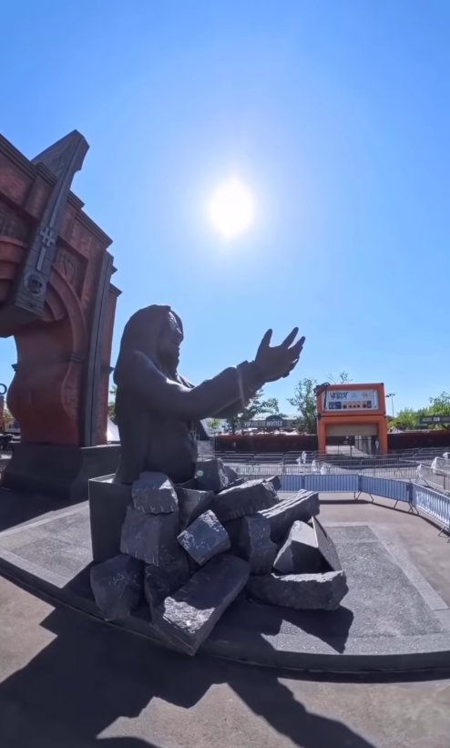 Seen in profile beneath the midday sun, Ozzy Osbourne's statue stretches its arms outward from a base of broken stone. Festival tents, barriers, and nearby buildings frame the tribute at Hellfest in Clisson, France.