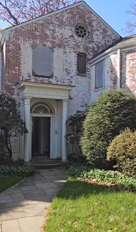 The front entrance of Richard Gere's former Connecticut estate featured a weathered brick facade, a columned portico, and mature landscaping surrounding a stone walkway.