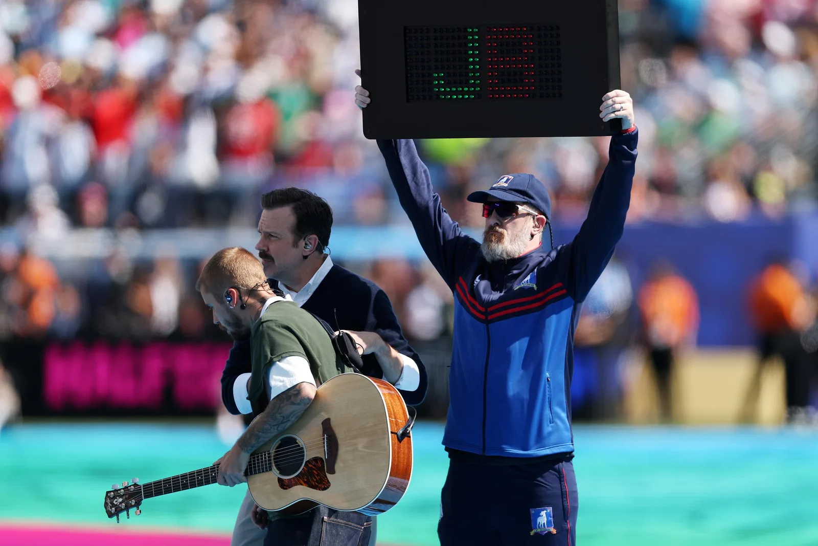 A stadium crew member holds an electronic board displaying a giant "J" and "US" during Justin Bieber's halftime appearance, adding a branded visual to the musician's low-energy acoustic interlude.
