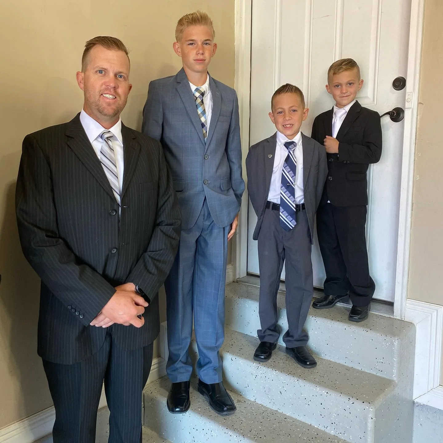 Spencer stands at the forefront with hands respectfully clasped, posing indoors with his three handsome sons, Reid, Thayne, and Gage, on a nearby stairway. The quartet looks unified and sharp in their tailored suits, representing a legacy of strength and unity.