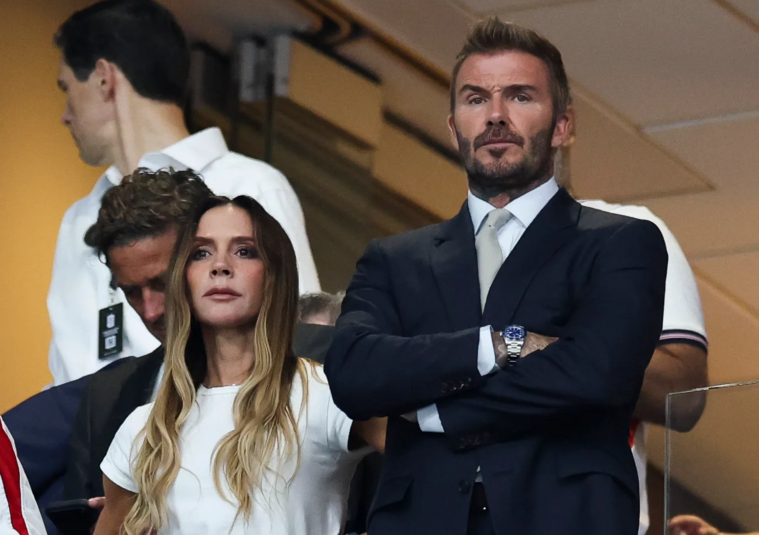 A focused Victoria Beckham, wearing a relaxed white top and jeans, stands beside David Beckham in his tailored navy suit as they watch football's biggest night with intent looks.
