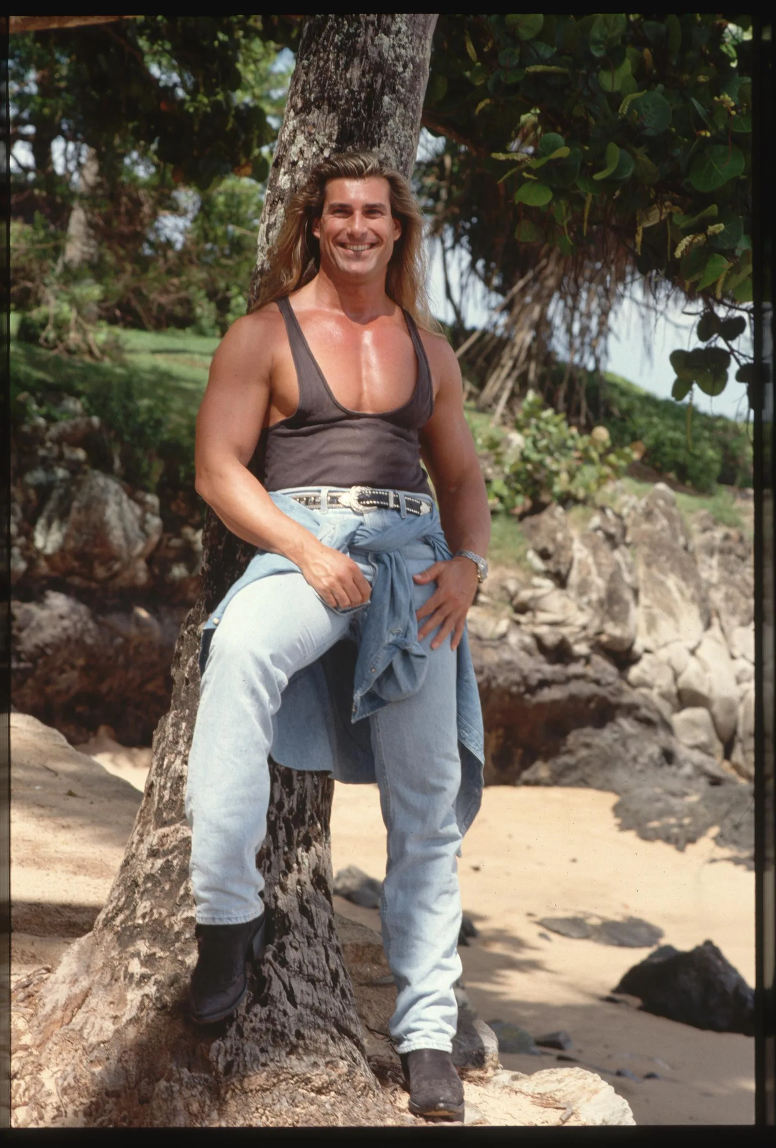 At first, he had no idea he was on any book covers. In this photo, the iconic romance book cover star wore a tank top and jeans while leaning against a tree in Hawaii on January 1, 1993.