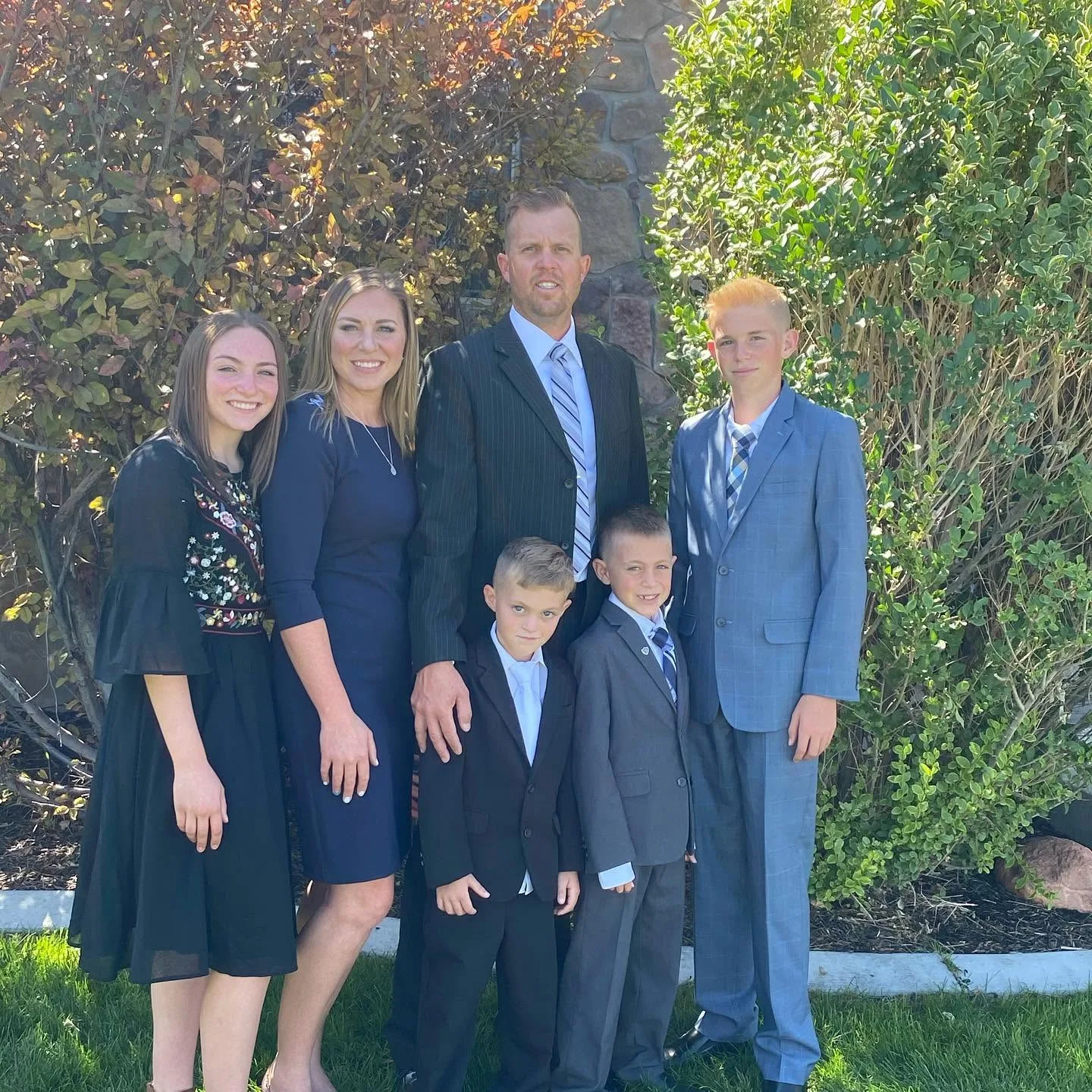 The entire Long family poses formally outdoors against a backdrop of green foliage and a stone wall. Katrina stands elegant in navy, Lydia in a detailed black dress, and Captain Spencer looks sharp alongside his three sons, Reid, Thayne, and Gage, all dressed in their sharpest suits.