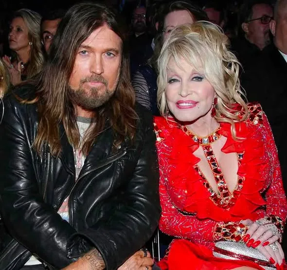 Dolly Parton, stunning in a bright red, embellished dress, sits alongside Billy Ray Cyrus, rocking his signature leather jacket and long hair, at a public event.